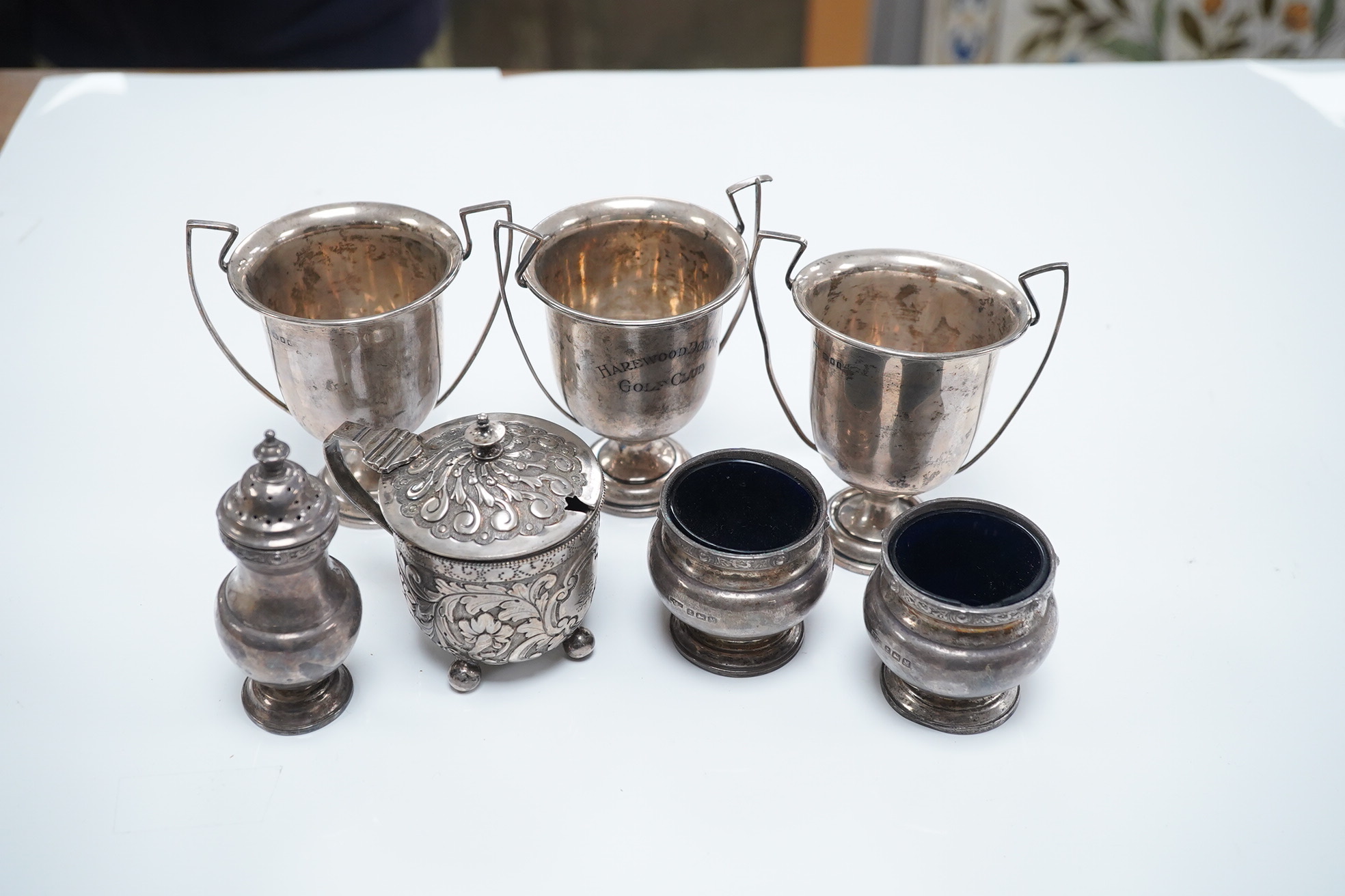 A Victorian repousse silver drum mustard, London, 1887, a silver three piece condiment set, Birmingham, 1936 and a set of three small trophy cups by Mappin & Webb, London, 1925/6. Condition - poor to fair